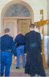 SSPX priest Fr. Michael McMahon being led by the men in blue through the vestibule, down the stairs, to talk things over with the authorities.  Notice the Crucifix over his right shoulder... very reminiscent of the Palace Guard,
leading Christ to His death. Will you follow Fr. Michael and take up your Cross as well?